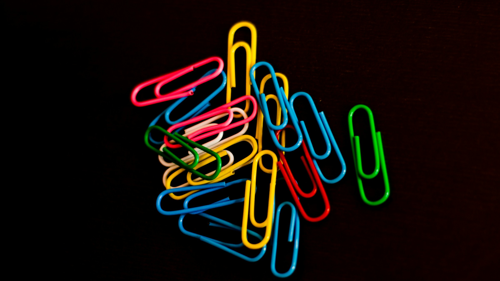 A pile of colourful paperclips on a black background, with one green clip set apart