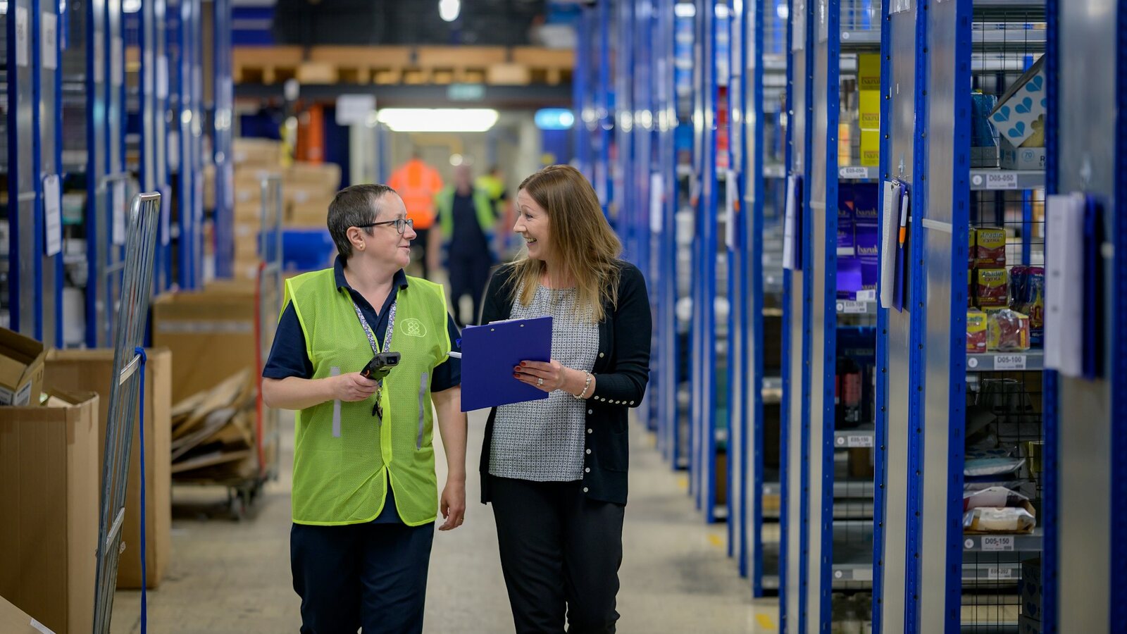 Two warehouse staff walking a long aisle of stocked shelving with a clipboard