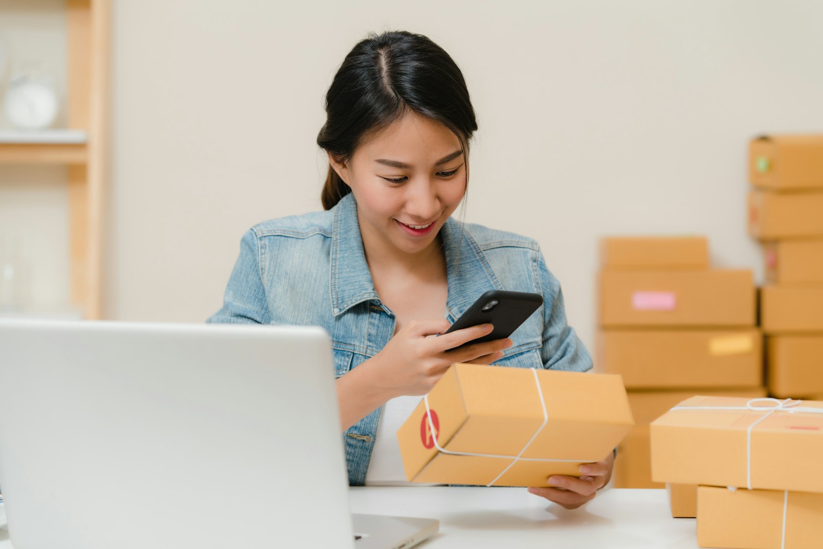 Woman at a home office scanning a code on a product with her phone, with a laptop and shipping boxes nearby