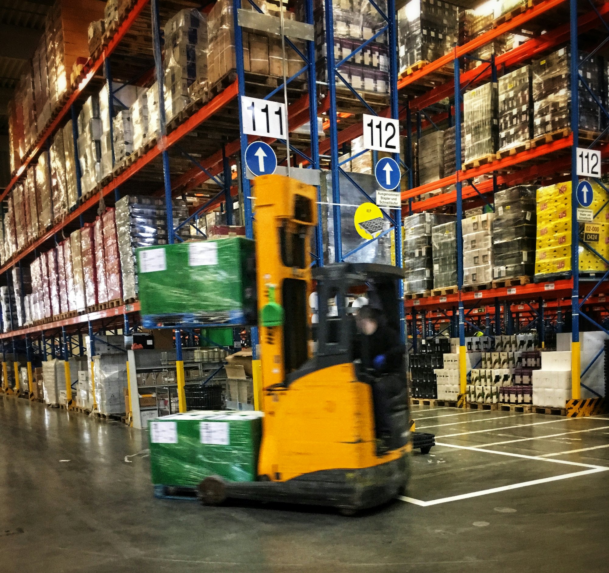 A forklift driving through a warehouse aisle lined with pallets on racking