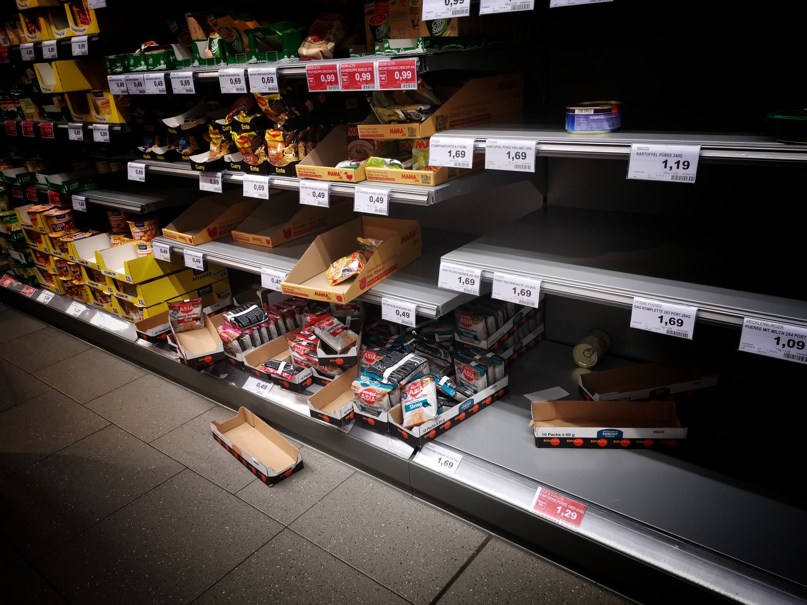 Supermarket shelves with some sections empty and others still stocked, with price tags and cardboard boxes