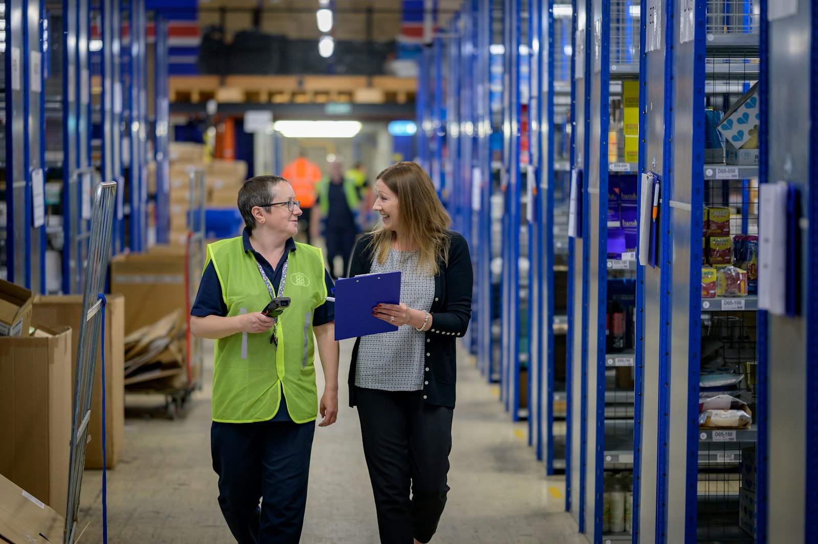 Two women in high-visibility vests walking along a warehouse aisle with blue shelving, one holding a clipboard